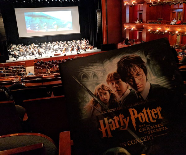 Concert hall with orchestra on the stage and a screen showing a Quidditch scene with the program book for Harry Potter and the Chamber of Secrets